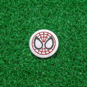 Op de afbeelding: Een ronde, zilverkleurige golfbalmarker met het ontwerp van het Spider-Man-masker. Het ontwerp bevat een rood webpatroon en zwarte oogcontouren op een witte achtergrond. De marker ligt op een groene kunstgras achtergrond.