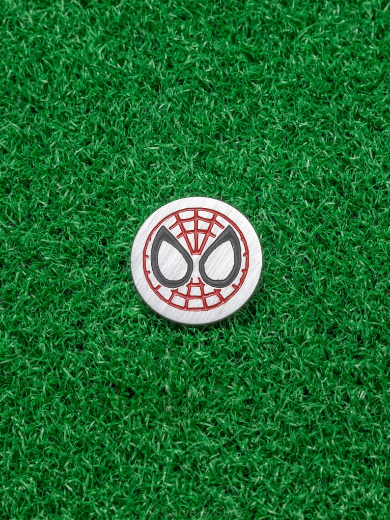 Op de afbeelding: Een ronde, zilverkleurige golfbalmarker met het ontwerp van het Spider-Man-masker. Het ontwerp bevat een rood webpatroon en zwarte oogcontouren op een witte achtergrond. De marker ligt op een groene kunstgras achtergrond.