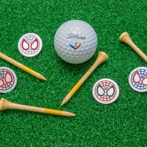 Op de afbeelding: Een golfbal, tees en balmarkers zijn gerangschikt op groen gras. De witte golfbal heeft het woord "Titleist" erop gedrukt. De tees zijn van hout met gele punten. De balmarkers hebben een Spider-Man-ontwerp.