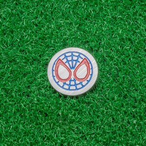 Op de afbeelding: Een ronde, zilverkleurige golfbalmarker met het iconische Spider-Man maskerontwerp. Het masker heeft een blauwe omtrek met rode ogen en een webpatroon. Geplaatst op een achtergrond van heldergroen kunstgras.