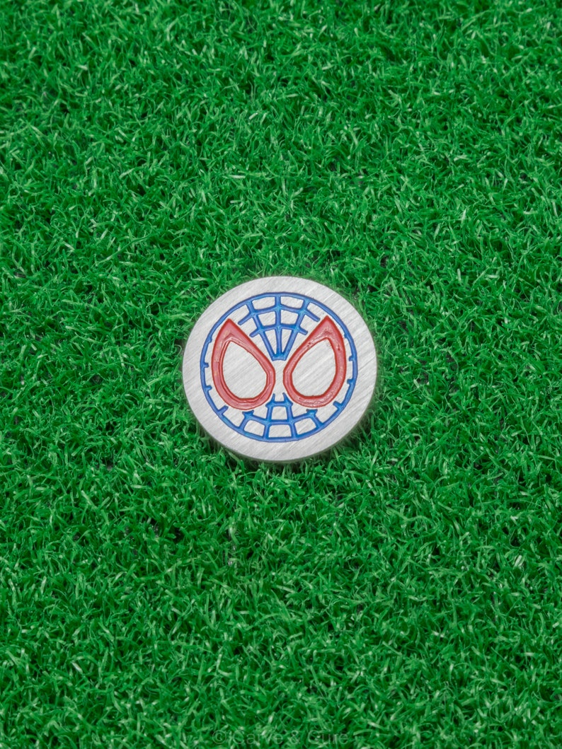Op de afbeelding: Een ronde, zilverkleurige golfbalmarker met het iconische Spider-Man maskerontwerp. Het masker heeft een blauwe omtrek met rode ogen en een webpatroon. Geplaatst op een achtergrond van heldergroen kunstgras.