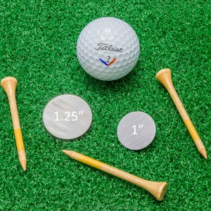 Op de afbeelding: Een golfbal, twee houten golf tees en twee zilverkleurige markers zijn gerangschikt op een groene ondergrond. De golfbal is wit met het woord "Titleist" erop gedrukt. De markers zijn gelabeld met "1.25" en "1".