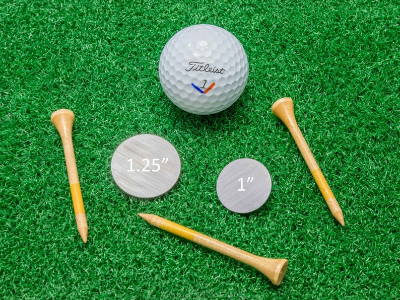 Op de afbeelding: Een golfbal, twee houten golf tees en twee zilverkleurige markers zijn gerangschikt op een groene ondergrond. De golfbal is wit met het woord "Titleist" erop gedrukt. De markers zijn gelabeld met "1.25" en "1".