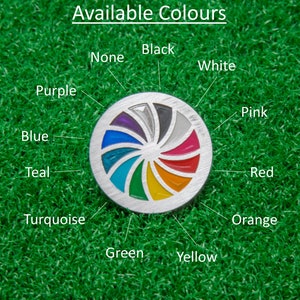 Op de afbeelding: Een zilverkleurige golfbalmarker met een kleurrijk spiraalontwerp, met kleuren als paars, blauw en rood. De tekst "Available Colours" staat bovenaan, met kleurnamen rond de marker.