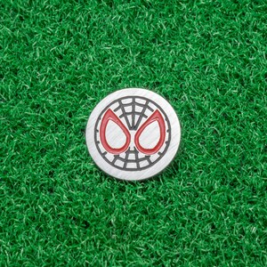 Op de afbeelding: Een ronde, zilverkleurige golfbalmarker met het iconische Spider-Man-maskerontwerp. De ogen zijn rood en het web is zwart. De marker ligt op een achtergrond van felgroen kunstgras.