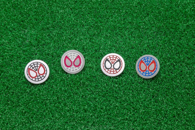 Op de afbeelding: Vier zilveren golfbalmarkers met het Spider-Man maskerontwerp in rood, zwart en blauw. De markers liggen op een groene kunstgras ondergrond.