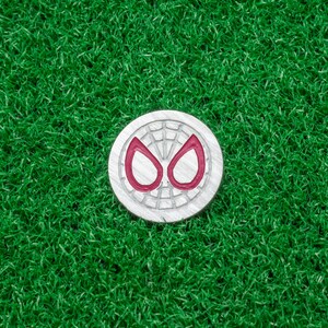 Op de afbeelding: Zilverkleurige golfbalmarker met een rood en zilver spinnenogen-ontwerp. De ronde marker ligt op een groene kunstgras ondergrond.