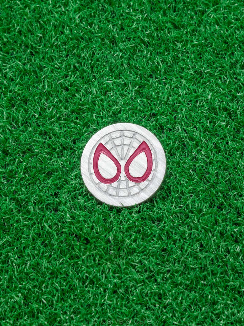 Op de afbeelding: Zilverkleurige golfbalmarker met een rood en zilver spinnenogen-ontwerp. De ronde marker ligt op een groene kunstgras ondergrond.