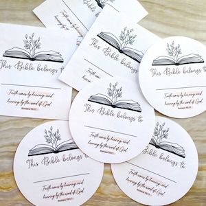 May include: A set of five white circular stickers with a black and white illustration of an open book with leaves on top. The text "This Bible belongs to" is printed on each sticker. The text "Faith comes by hearing and hearing by the word of God. Romans 10:17" is printed on four of the stickers.