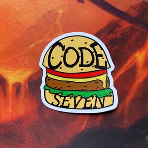 May include: A yellow and brown cartoon burger sticker with the words "CODE SEVEN" on it.
