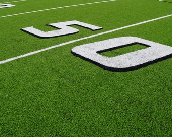 Pode incluir: Um campo de futebol de relva artificial verde com linhas de jarda brancas e os números 50 pintados no campo.