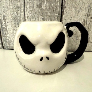 Può includere: Una tazza in ceramica bianca a forma di volto di Jack Skellington da The Nightmare Before Christmas. La tazza ha grandi orbite nere, una bocca cucita e un manico nero. Un oggetto unico per i fan del film.
