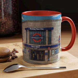 Puede incluir: Taza de cerámica con interior y asa rojos, con un diseño de estación de metro de Londres. La taza tiene el texto "Ruining Your Day" sobre el logo de Underground y "Since 1863" debajo. Una cuchara y un macaron están sobre una superficie de madera.