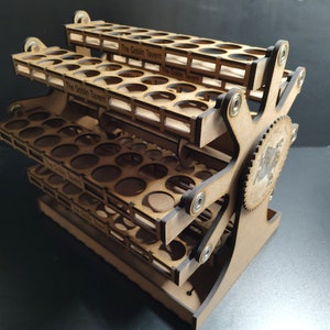 May include: A wooden paint rack with multiple tiers, each designed to hold paint bottles. The rack has a gear-like mechanism on the side and the text "The Goblin Tavern" is visible on the front of each tier. The rack is made of light brown wood.