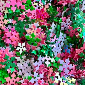 May include: A close-up view of a pile of snowflake-shaped confetti. The confetti is a mix of colors, including pink, green, white, and iridescent. The snowflakes are scattered and overlapping, creating a textured and festive appearance.
