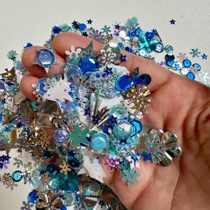 May include: A collection of loose craft sequins and glitter pieces in shades of blue, silver, and white. The assortment includes various shapes such as snowflakes, stars, triangles, and circular discs with metallic, iridescent, and matte finishes.