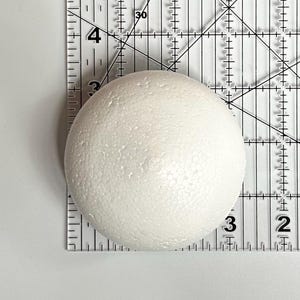 May include: A white styrofoam ball, approximately 3 inches in diameter, is shown on a clear ruler.