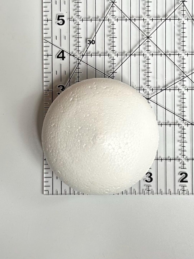 May include: A white styrofoam ball, approximately 3 inches in diameter, is shown on a clear ruler.