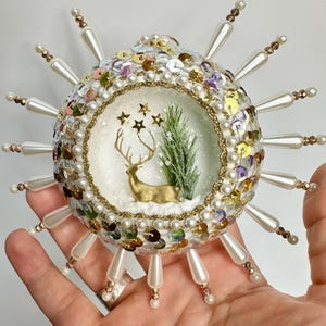 May include: A white Christmas ornament with a gold deer and a small green tree inside. The ornament is covered in sequins and pearls and has a white beaded fringe around the edge.