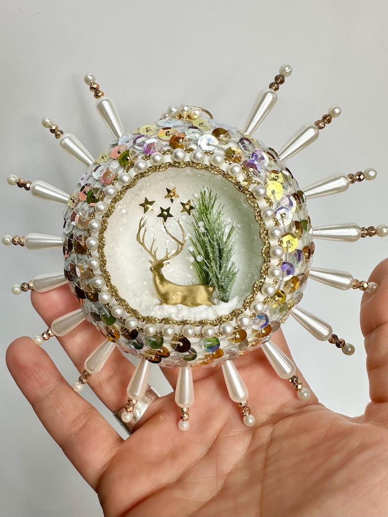 May include: A white Christmas ornament with a gold deer and a small green tree inside. The ornament is covered in sequins and pearls and has a white beaded fringe around the edge.