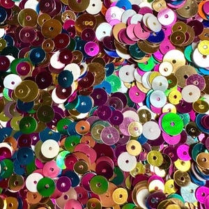 May include: A close-up shot of a pile of colorful sequins. The sequins are round with a hole in the center and come in various colors, including gold, pink, green, blue, and white. The sequins are scattered and overlapping, creating a textured surface.