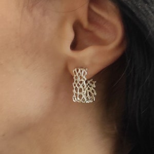 May include: A pair of silver hoop earrings with a delicate, intricate design. The earrings are made of a woven metal mesh.