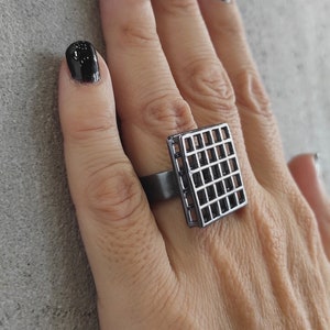 May include: A black square ring with a grid pattern. The ring is worn on the middle finger of a hand with black nail polish.