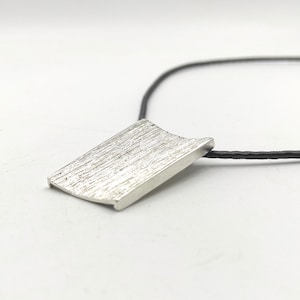 May include: A silver pendant necklace with a textured rectangular pendant on a black cord.