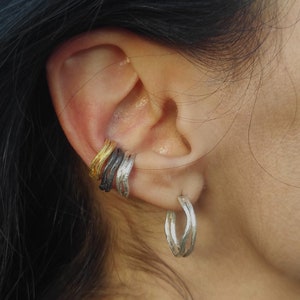 May include: A person wearing a variety of silver and gold hoop earrings. The earrings are stacked on the earlobe and are made of thin, twisted metal.