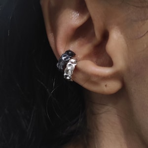 Op de afbeelding: Een paar oormanchetten, een zwart en een zilver, gedragen op het bovenste deel van het oor.