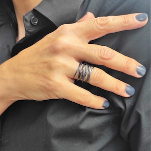 Könnte beinhalten: Ein schwarzer Metallring mit einem gedrehten Design. Der Ring wird am Zeigefinger einer Hand mit dunklem Nagellack getragen.