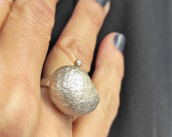 May include: A silver ring with a textured, rounded top and a small, clear stone on top. The ring is worn on a person's finger.