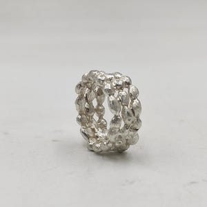 Peut inclure: Une bague en argent au design texturé et organique. La bague présente trois rangées d'éléments ovales reliés, créant un bijou unique et accrocheur. La bague est présentée sur un fond blanc uni.