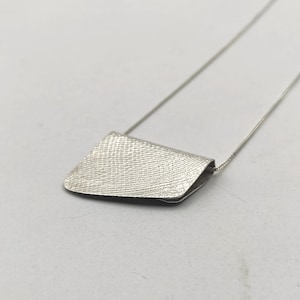 Op de afbeelding: Een zilveren ketting met een getextureerde, geometrische hanger. De hanger is gevormd als een klein, plat tasje.