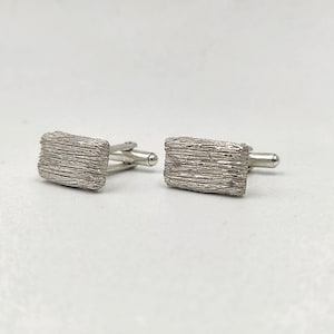Puede incluir: Un par de gemelos de plata con un acabado texturizado cepillado. Los gemelos son de forma rectangular y tienen un diseño sencillo.