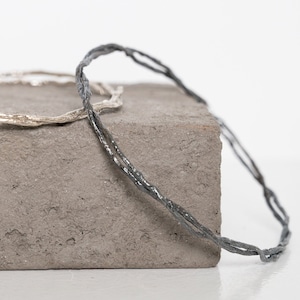 Op de afbeelding: Twee zilverkleurige armbanden. Eén is donkergrijs, de andere lichter zilver. De armbanden hebben een textuur, organisch ontwerp. Ze rusten op een grijs, rechthoekig blok tegen een witte achtergrond.
