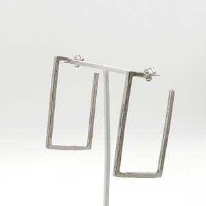 May include: A pair of silver square hoop earrings. The earrings are made of a textured metal and have a simple, modern design.