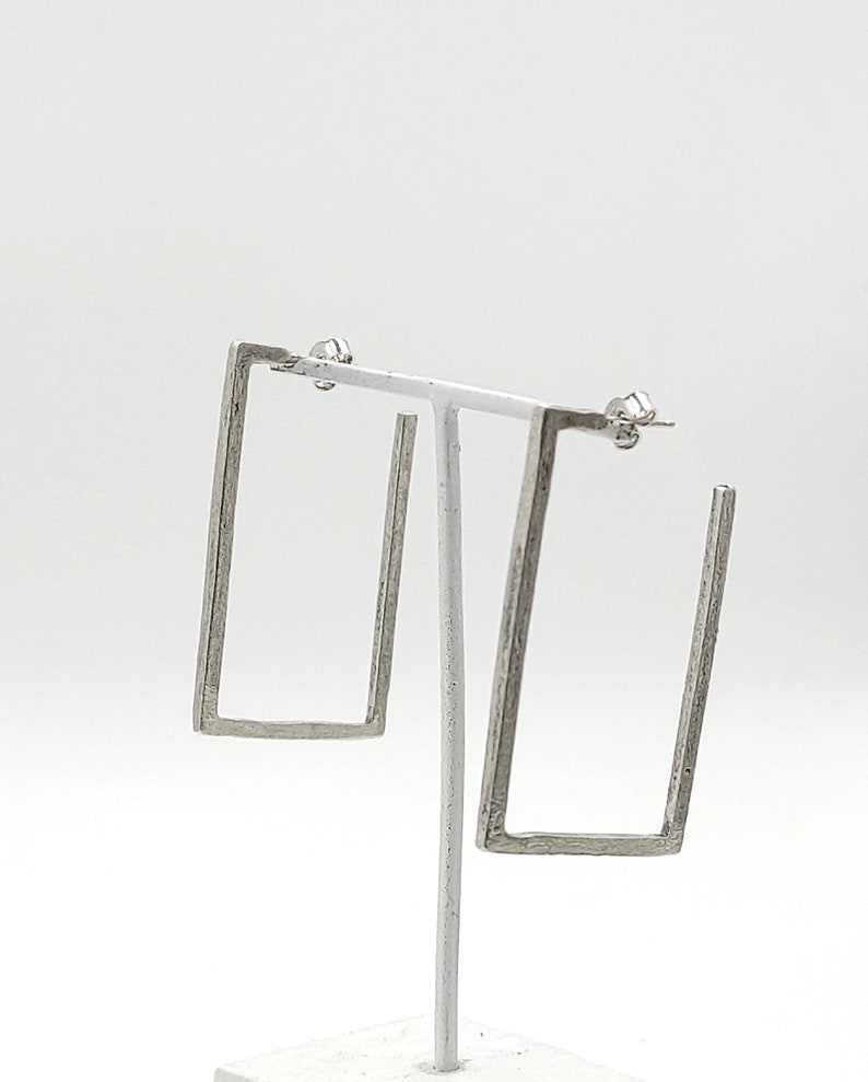 May include: A pair of silver square hoop earrings. The earrings are made of a textured metal and have a simple, modern design.