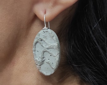 May include: A pair of gray concrete earrings with a textured surface and a silver hook. The earrings are oval shaped and have a subtle pattern.