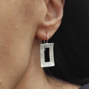 Könnte beinhalten: Silberne Ohrhänger mit einem strukturierten rechteckigen Design. Das Rechteck hat einen Ausschnitt in der Mitte, der eine rahmenartige Form erzeugt.