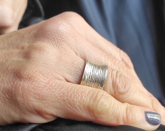 May include: A silver ring with a textured, layered design. The ring is worn on the ring finger of a person's hand.