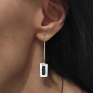 Könnte beinhalten: Silberne Ohrringe mit einem schwarzen rechteckigen Anhänger. Der Anhänger hat einen strukturierten silbernen Rahmen.