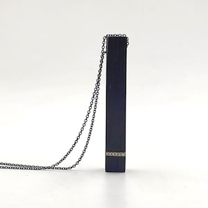 Puede incluir: Un collar negro con un colgante rectangular negro que presenta una fila de pequeños diamantes. La cadena es negra y tiene un eslabón delicado.