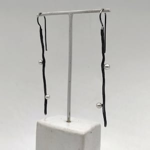 Puede incluir: Un par de pendientes alargados de color gris oscuro con un diseño similar a una rama. Cada pendiente presenta tres pequeñas cuentas redondas de color plateado. Los pendientes cuelgan de un gancho simple y se exhiben sobre un fondo blanco.