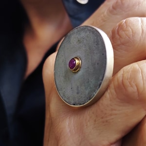 Puede incluir: Un gran anillo plateado con un centro de piedra gris circular. Un pequeño engaste dorado sostiene una vibrante gema roja. El anillo se lleva en una mano, con una camisa azul oscuro en el fondo.