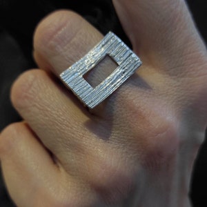 Può includere: Un anello d'argento con un designo rettangolare testurizzato che presenta un ritaglio al centro. L'anello è indossato su un dito.