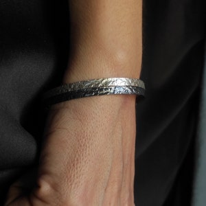 May include: Two bracelets on a person's wrist. The top bracelet is silver with a textured surface. The bottom bracelet is black leather.