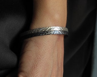 May include: Two bracelets on a person's wrist. The top bracelet is silver with a textured surface. The bottom bracelet is black leather.