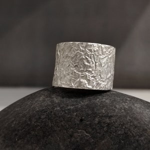 May include: A silver ring with a textured surface. The ring is sitting on a dark grey stone.