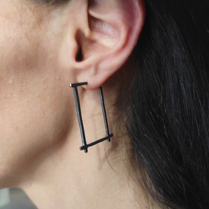 May include: A close-up of a person's ear wearing a pair of black rectangular earrings. The earrings are made of thin, dark metal bars, forming a geometric shape. The earrings are attached to the earlobe.